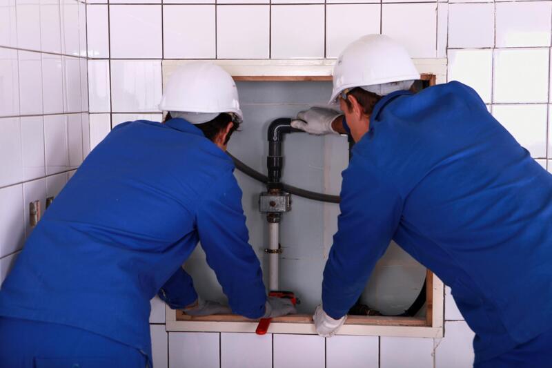 two plumbers working on plumbing behind a wall